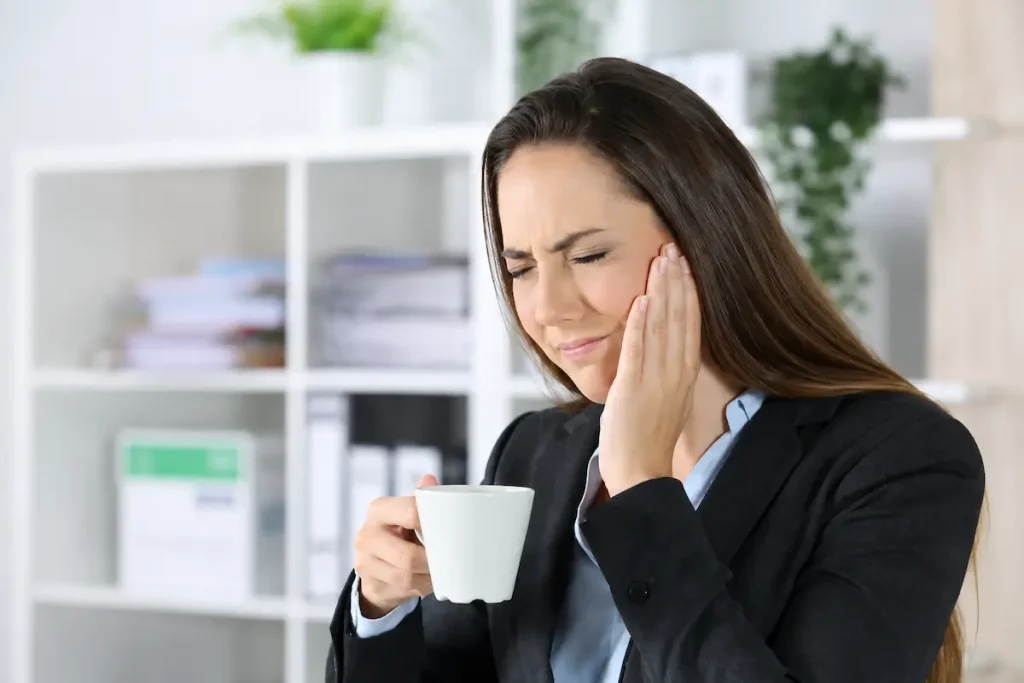 Acupuncture for TMJ Disorders: A Setauket Patient's Evidence-Based Guide 1 Woman holding her jaw in pain while drinking coffee in an office, representing acupuncture for TMJ treatment and natural jaw pain relief.